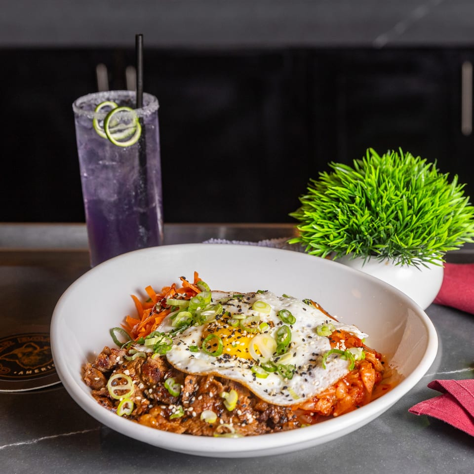 A white bowl of bulgogi, kimchi, carrots, and a fried egg, with a purple drink and plant.
