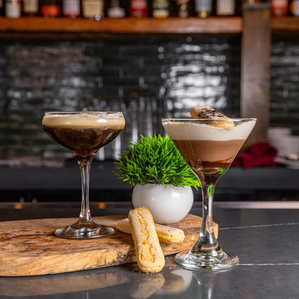 Two dessert cocktails, one dark and foamy, the other layered with cream, on a bar.