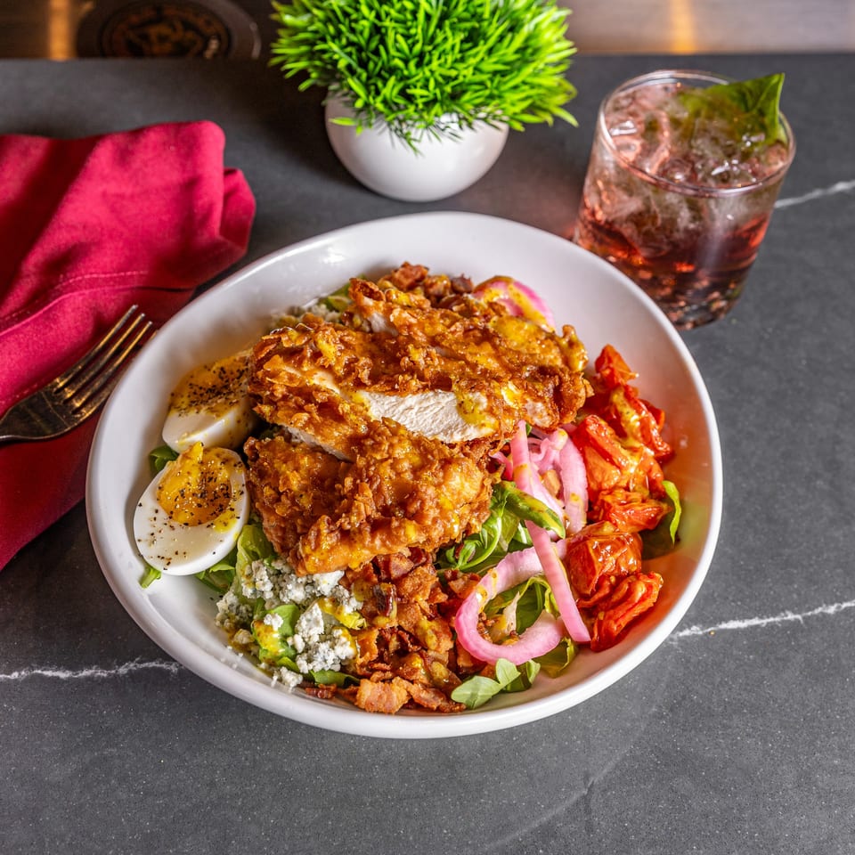 Crispy fried chicken salad with hard-boiled eggs, bacon, tomatoes, and pickled onions, with a drink.