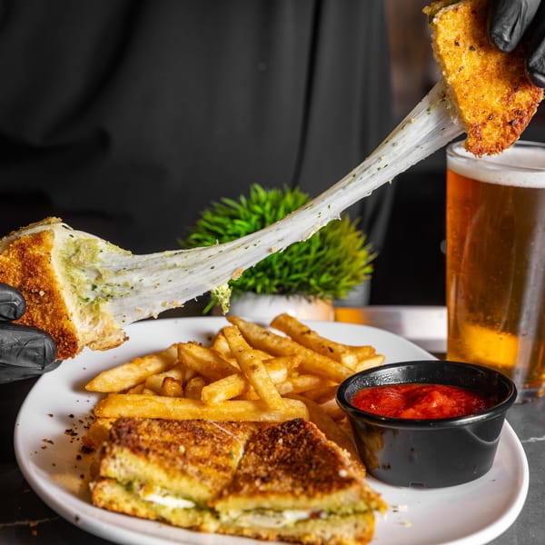 Hand in black glove pulling apart a pesto grilled cheese with melted cheese stretching, served with fries and marinara.