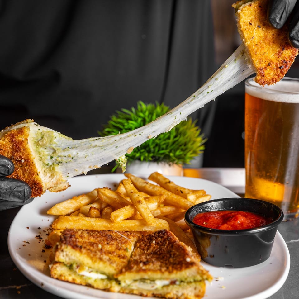 Hand in black glove pulling apart a pesto grilled cheese with melted cheese stretching, served with fries and marinara.