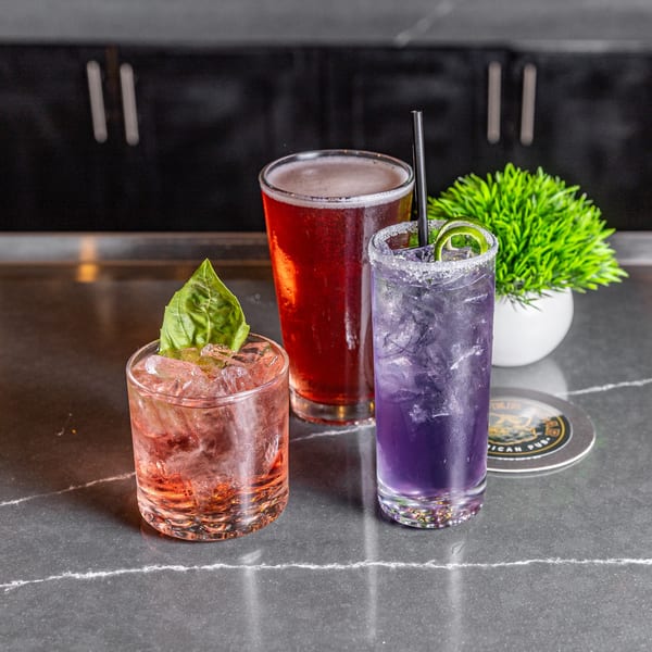 Three drinks on a dark countertop: a red cocktail with basil, a beer, and a purple cocktail.