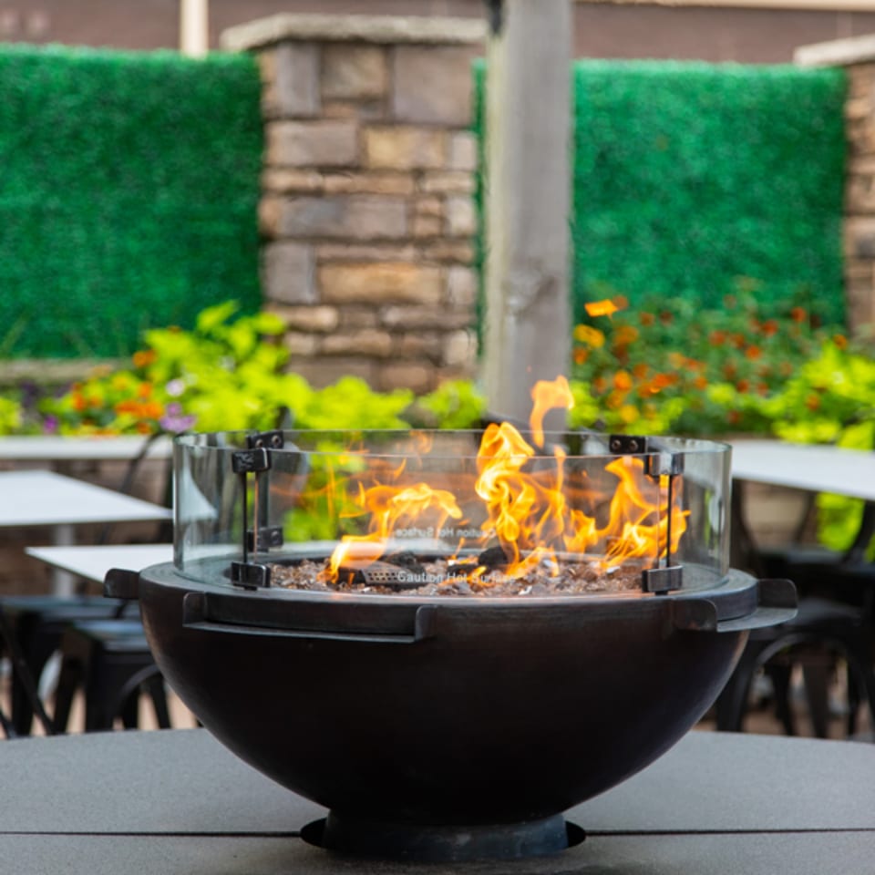 A black outdoor fire pit with a glass shield, flames, and green foliage in the background.
