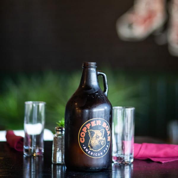 A Copper Boar American Pub growler sits on a dark table with glasses and a pink napkin.