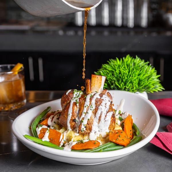 Gravy pouring over a braised pork shank with mashed potatoes, green beans, and carrots.