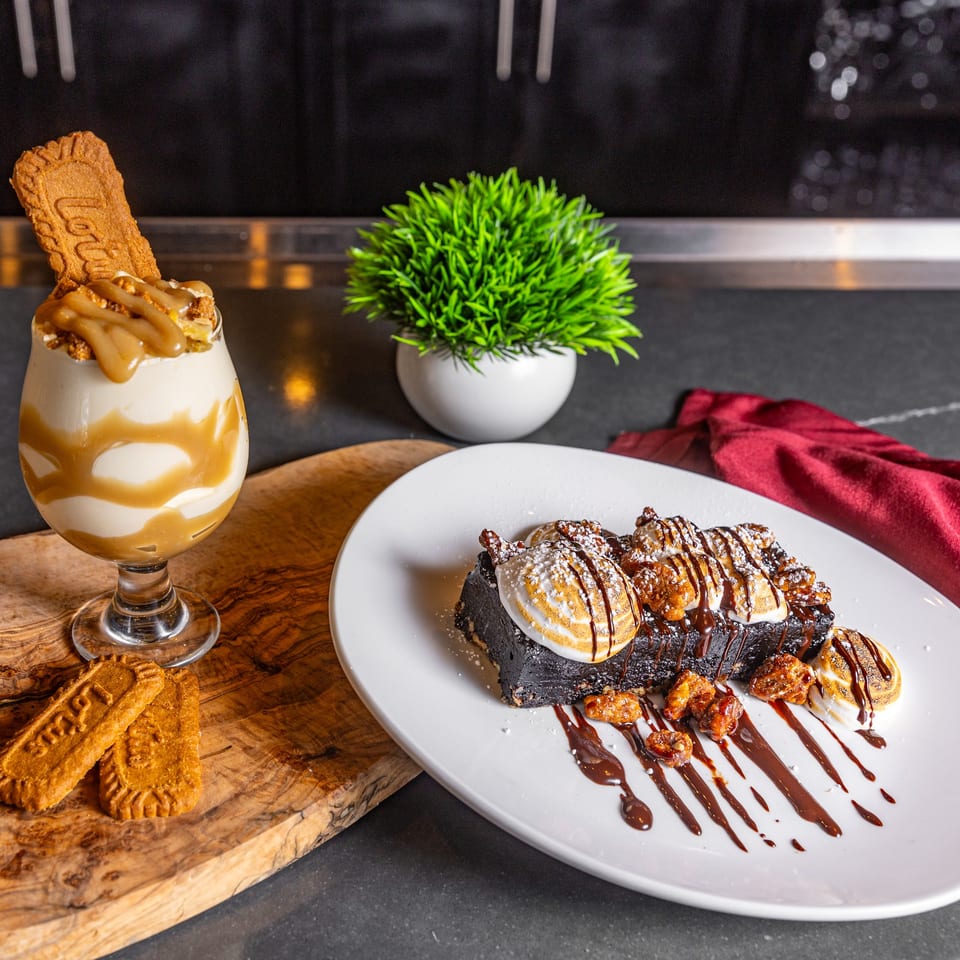 A rich chocolate brownie with toasted marshmallows, candied nuts, and chocolate drizzle, next to a caramel milkshake.