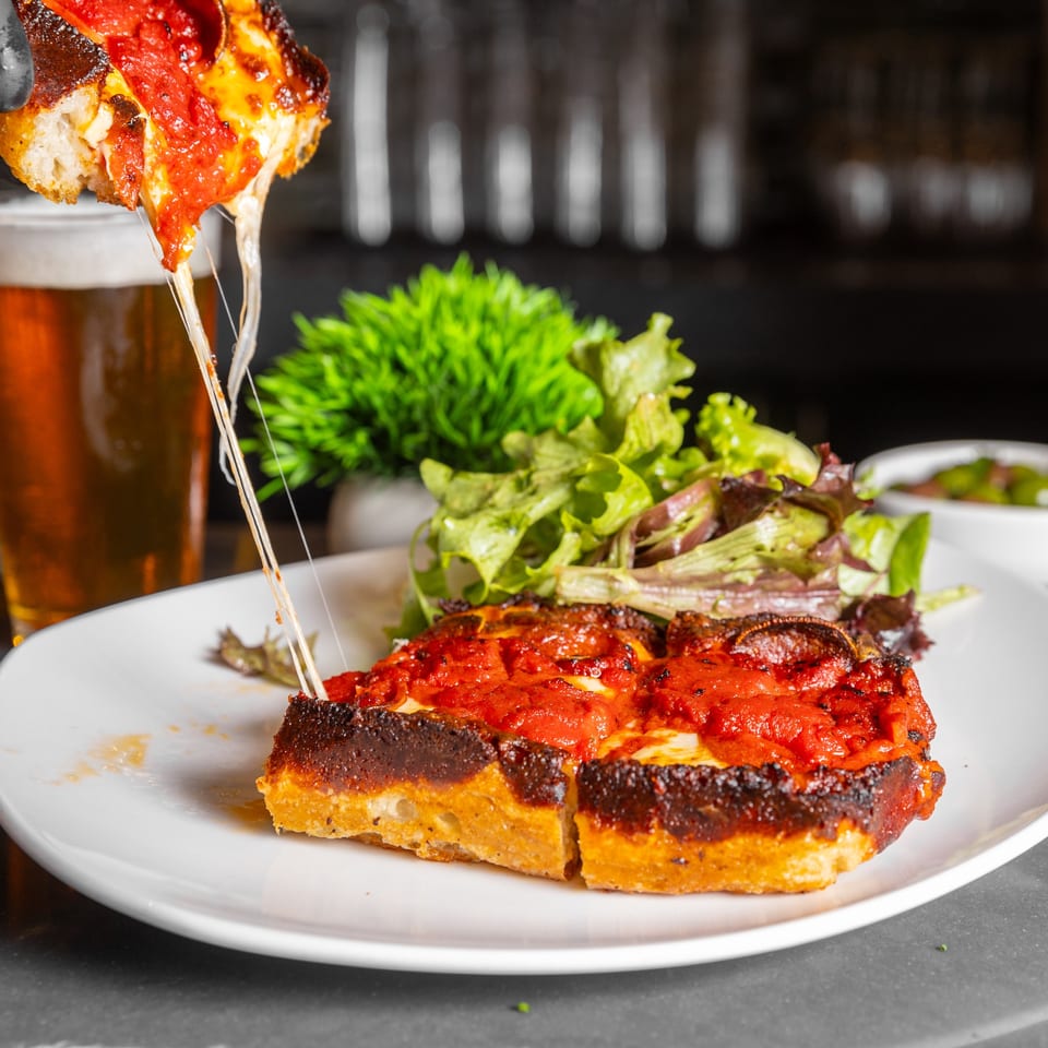 Deep dish pizza slice with melted cheese pull, side salad, and beer at Copper Boar American Pub.