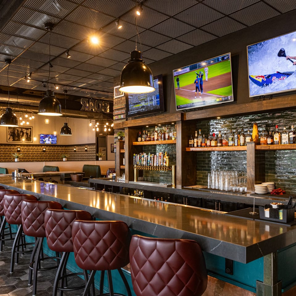 A long, well-lit bar with red leather stools, multiple beer taps, liquor bottles, and TVs showing sports.